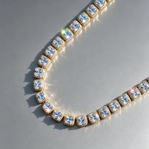 10MM Cushion Clustered Tennis Chain in Gold lay‑flat angled product shot on seamless grey reflective surface, sunlit natural lighting, brilliant starburst sparkles and rainbow light flares, hand‑set cushion halo VVS simulated diamond links, thick gold‑plated hip‑hop bling necklace, unisex statement jewelry, realistic studio product photography RDRIP JEWELRY