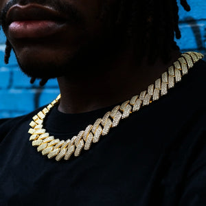 Close-up shot of Black male model with neat dreadlocks and neatly trimmed short beard wearing 19mm full iced out prong set rectangular baguette cuban link chain, premium hip hop bling necklace with flawless VVS simulated diamond stones, solid copper base with polished 18K gold finish, model dressed in plain black crew neck tee, vibrant blue graffiti brick wall street background, heavy wide men's statement hiphop jewelry RDRIP JEWELRY