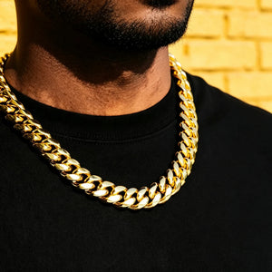 Close-up shot of men wearing chunky 18mm yellow gold plated Miami Cuban link choker necklace, heavy thick polished curb chain hip hop streetwear jewelry, short trimmed beard, clean solid black plain tee, warm yellow brick wall background, shiny reflective gold finish oversized statement Cuban chain for men, tight interlocking links box clasp heavyweight stacking necklace, luxury street hip-hop accessory daily wear layering chain RDRIP JEWELRY since 2016