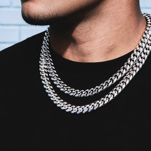 Close-up lifestyle shot of clean jawline with light stubble male model wearing double layered 12.2mm non-stone 14k white gold plated Miami Cuban link choker with secure box lock clasp, plain solid black crew neck t-shirt, soft pale light blue textured brick wall background, thick tightly interlocking highly polished curb links, cool reflective silver-white anti-tarnish plating, heavy weight uniced plain stackable hip hop necklace for men streetwear daily layering, minimalist premium men bling jewelry