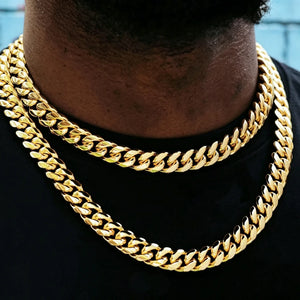 Extreme close-up shot of 12.2mm wide 18k gold plated Miami Cuban link choker with sturdy box clasp, two layered stacked heavy curb gold chains worn on Black male neck, soft minimal light blue wall background with subtle faint street graffiti details, tight polished interlocking cuban links, bright reflective anti-tarnish gold finish, thick hip hop layering necklace for men streetwear, bold icy luxury street jewelry closeup product photo, stackable gold curb chain everyday fashion accessory RDRIP JEWELRY
