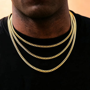 Front close-up portrait of black male model with trimmed short stubble beard wearing three layered stackable 5mm Miami Cuban Link Chain in 18k gold plated, polished glossy solid cuban link necklace for men hip hop streetwear, shot on soft light peach background, flexible tangle-free construction, available lengths 18 20 24 30 inch with weight tolerance ±5 grams, anti-tarnish durable 18k gold plating, can be worn alone or matched with custom hip hop pendants, classic urban layering jewelry gift for men