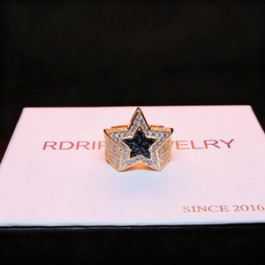 Flat‑lay front view of men’s gold‑plated iced out double pentagram star hip hop ring, hand‑set AAA white cubic zirconia stones surrounding solid black CZ star center, heavy wide micro pave band bling statement ring, luxury rap street fashion jewelry for party wear and men’s gift collection from RDRIP JEWELRY