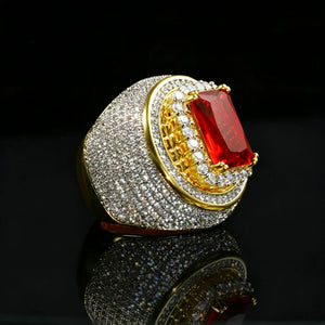 Luxury product photograph of heavy two‑tone 18k gold plated men hip‑hop champion ring, large radiant‑cut red ruby center stone secured by gold prongs, multi‑layer halo design with full micro pave hand‑set cubic zirconia stones, thick wide heavy band, brilliant icy bling shine, displayed on glossy black mirror reflective surface, dark studio background, premium rapper statement jewelry RDRIP JEWELRY