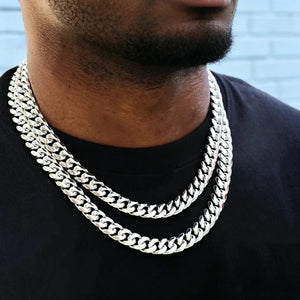 Close-up portrait of Black male wearing two layered 12mm non-stone 14k white gold plated Miami Cuban link choker with secure box clasp, neatly trimmed shortened beard, plain black crew neck t-shirt, soft light blue textured brick wall background, thick tight polished interlocking curb links, bright cool silver-white anti-tarnish finish, heavy weight stackable hip hop streetwear necklace for men, plain uniced-out luxury jewelry layering outfit shot RDRIP JEWELRY SINCE 2016