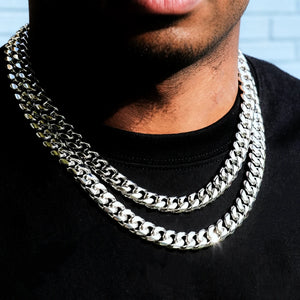 Close-up natural light shot of layered 12.2mm non-stone 14k white gold plated Miami Cuban link choker with secure box lock clasp, Black male model with neatly trimmed short beard wearing plain black crew neck shirt, soft pale light blue textured brick wall background, thick tightly polished interlocking curb links, cool reflective silver-white anti-tarnish finish, heavy weight uniced plain hip hop stackable necklace for men streetwear daily layering fashion, premium men bling jewelry product photo RDRIP JEW