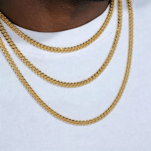 Product display close-up shot of three layered stackable 5mm Miami Cuban Link Chain with polished 18k gold plated finish, fitted on dark brown Black male neck mannequin wearing plain clean white crew neck t-shirt, shiny reflective gold cuban links with smooth flexible tangle-free construction, available necklace lengths 18 inch, 20 inch, 24 inch, 30 inch, long-lasting anti-tarnish gold plating resistant to fading and oxidation, versatile stackable layering chain that can be worn alone or matched with custom