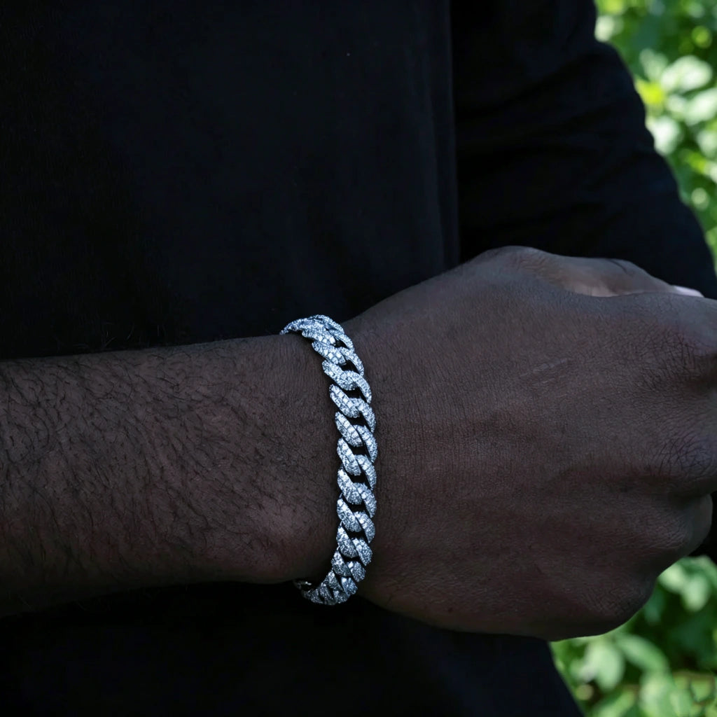 Front wrist close up of men's 10.5mm full iced out white gold plated cuban link bracelet, custom molded copper hip hop wrist jewelry with hand-set prong-lifted flawless VVS simulated diamonds, dark skin male model wearing icy miami cuban bracelet, natural lush green foliage plant background, heavy bling streetwear rapper accessory, available in 7", 8", 9" lengths, weight varies +/-5 grams, premium shiny diamond encrusted bracelet gift for hip hop fans, tarnish resistant fashion statement wrist chain, RDRIP 