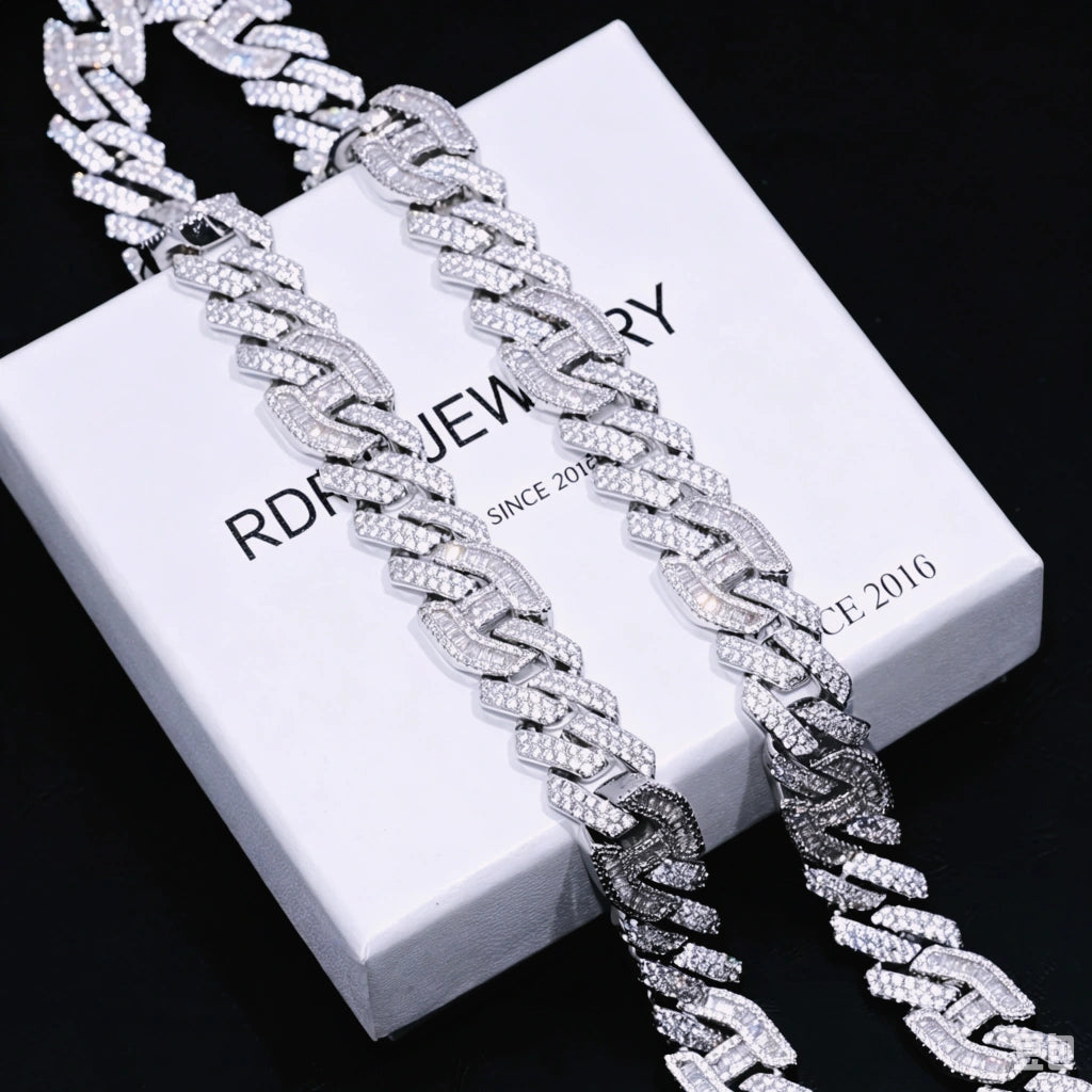 Angled partial close‑up shot of two 15mm prong‑set baguette mixed‑stone iced cuban curb chains in white gold finish, resting atop branded RDRIP JEWELRY gift box printed with SINCE 2016, hand‑set VVS simulated baguette cut and round diamonds, heavy full‑iced curb link details, dark textured black surface background, premium men hip‑hop streetwear bling statement necklace RDRIP JEWELRY
