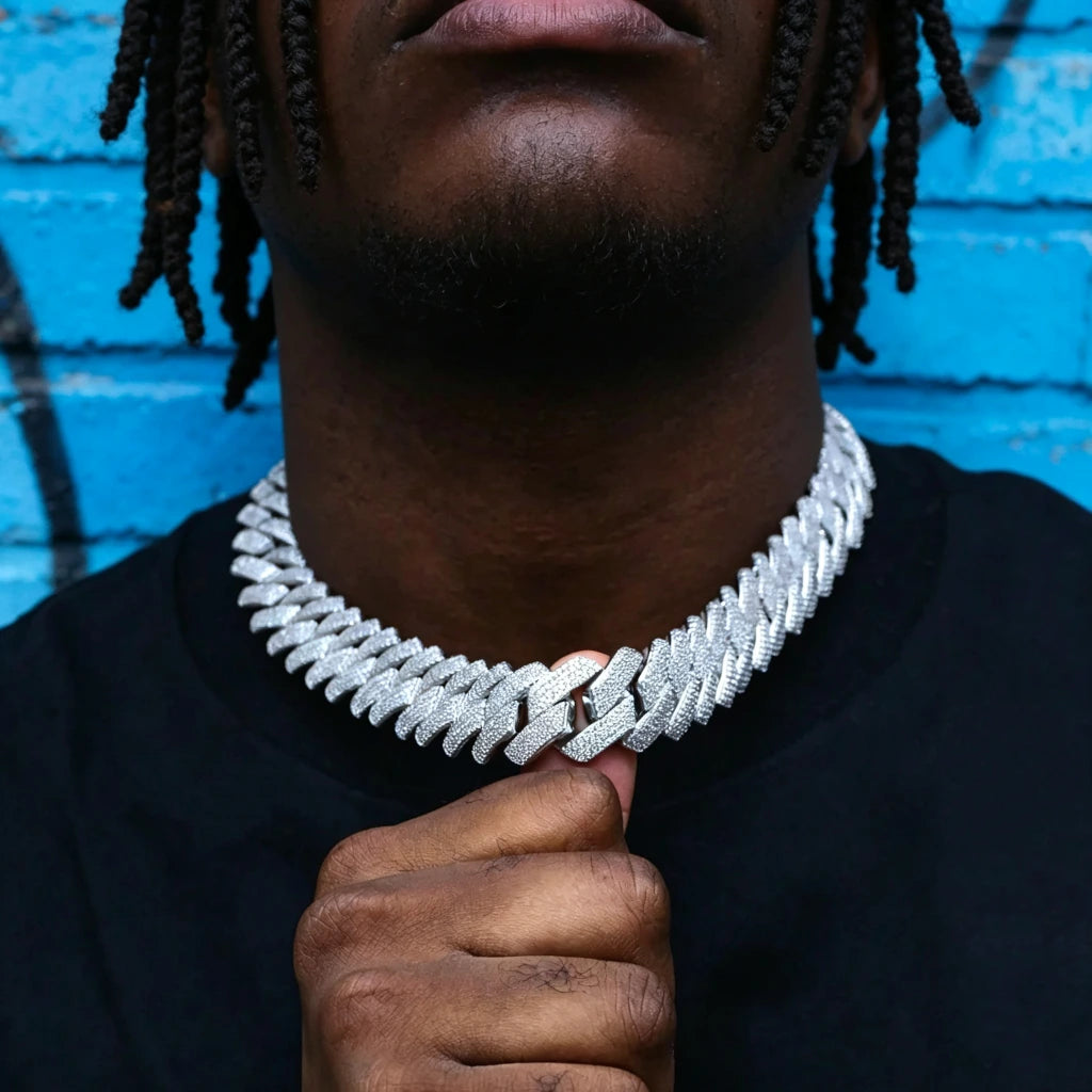 Close-up street style shot of Black male model with neat dreadlocks and neatly trimmed short beard holding the clasp of 19mm full iced out prong set rectangular baguette cuban link chain, polished 18K white gold plated copper hip hop necklace covered with flawless VVS simulated diamond stones, model wearing plain black crew neck shirt, vibrant blue graffiti painted brick wall background, heavy wide men statement bling jewelry detail shot RDRIP JEWELRY
