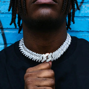 Close-up street style shot of Black male model with neat dreadlocks and neatly trimmed short beard holding the clasp of 19mm full iced out prong set rectangular baguette cuban link chain, polished 18K white gold plated copper hip hop necklace covered with flawless VVS simulated diamond stones, model wearing plain black crew neck shirt, vibrant blue graffiti painted brick wall background, heavy wide men statement bling jewelry detail shot RDRIP JEWELRY
