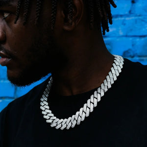 Men wearing 19mm full iced out prong set baguette cuban link chain necklace, thick wide hip hop bling cuban choker with fully paved rectangular VVS flawless simulated diamond stones, copper base 18k white gold finish, male model with short trimmed beard and long dreadlocks, shot against bright blue graffiti brick wall, trendy street hiphop men jewelry close up neck shot RDRIP JEWELRY
