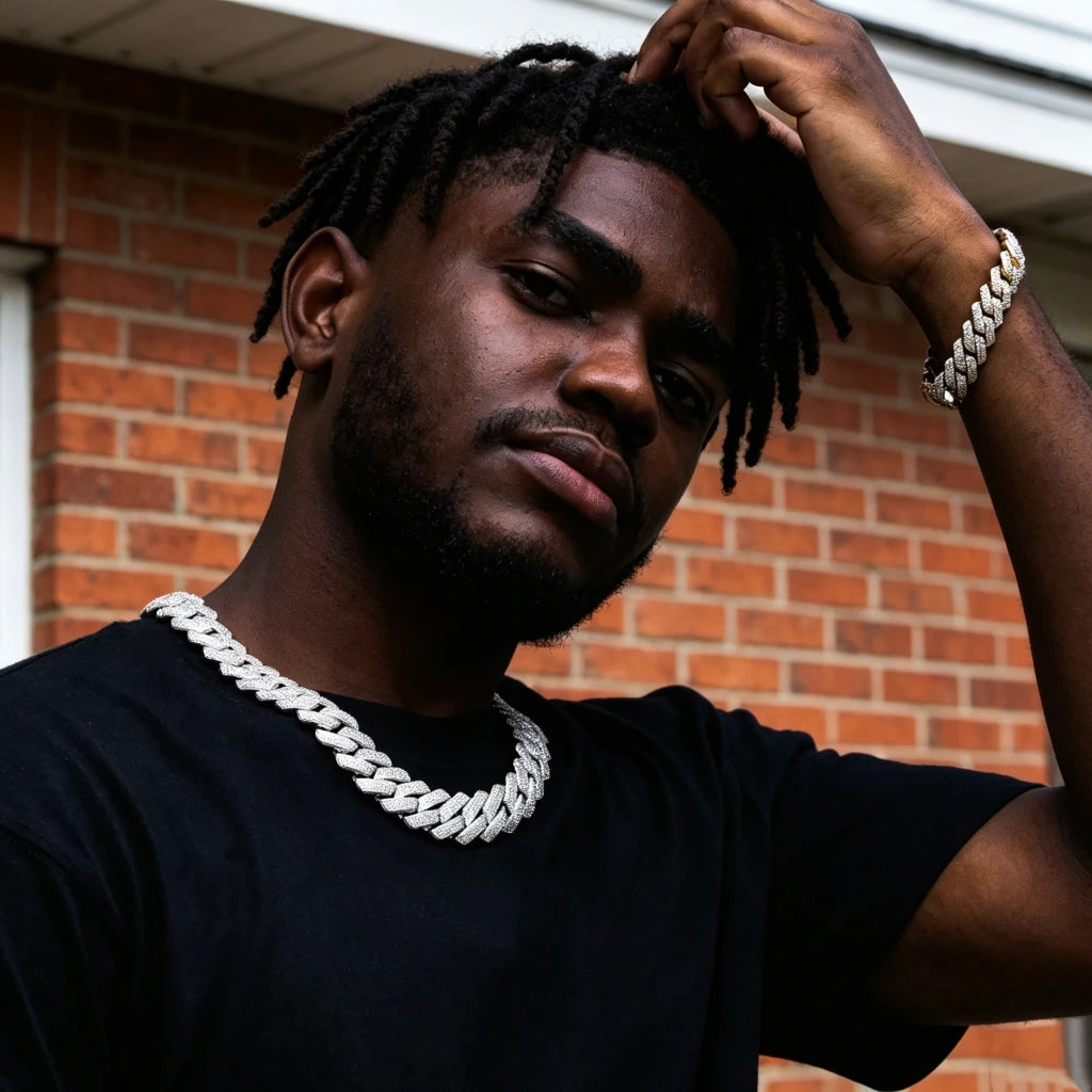 Street portrait shot of Black male model with neat long dreadlocks and neatly trimmed short beard wearing 19mm full iced prong set rectangular baguette cuban link necklace in polished 18K white gold finish, matching 18k gold fully iced baguette cuban bracelet on wrist, jewelry embedded with flawless VVS simulated diamond stones, solid copper base hand-set stones, model dressed in plain black short sleeve tee, one hand resting on hair, backdrop of rustic red brick house wall with faint graffiti, trendy 