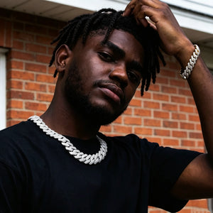 Street portrait shot of Black male model with neat long dreadlocks and neatly trimmed short beard wearing 19mm full iced prong set rectangular baguette cuban link necklace in polished 18K white gold finish, matching 18k gold fully iced baguette cuban bracelet on wrist, jewelry embedded with flawless VVS simulated diamond stones, solid copper base hand-set stones, model dressed in plain black short sleeve tee, one hand resting on hair, backdrop of rustic red brick house wall with faint graffiti, trendy 
