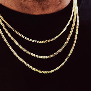 3mm 18k Gold Plated Italian Franco Link Chain layered stacked necklace for men and women, three size layered gold Franco chains worn by Black male model with short stubble beard, fitted solid plain black cotton t-shirt, shot against vivid red graffiti street art wall background, polished tangle-resistant flexible Franco link jewelry, shiny durable tarnish-free gold hip hop bling chain, compatible with custom hip hop pendants, minimalist everyday street fashion stacking necklace, premium unisex statement str