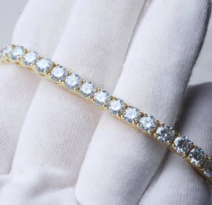 Bright studio close-up photograph of 4mm round brilliant cut D color VVS1 clarity moissanite tennis bracelet, solid S925 sterling silver base plated with thick 18K yellow gold, every clear moissanite stone securely hand prong set by skilled craftsmen, bracelet gently held by soft white cotton jewelry gloves on light neutral background, vivid rainbow sparkle and intense fire light reflecting from each flawless moissanite gemstone, unisex iced out hip hop luxury wrist chain for men and women, perfect for dail