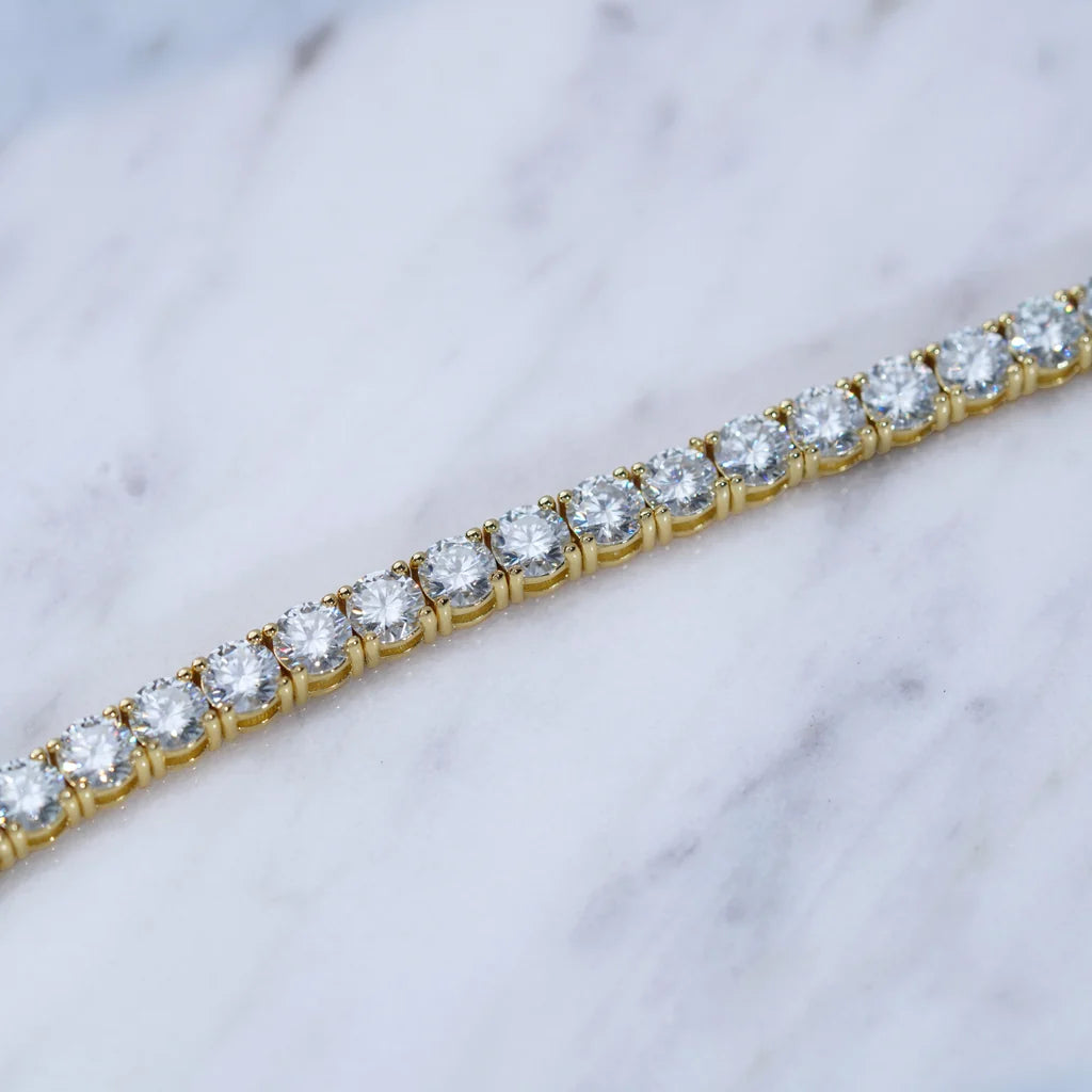 Horizontal close-up detail shot of 4mm round brilliant D color VVS1 clarity moissanite tennis bracelet, solid S925 sterling silver core with thick 18K yellow gold plating, each clear moissanite gem hand prong set with polished gold prongs, placed on clean white grey marble texture surface, bright natural lighting highlighting outstanding sparkle, fire and clarity of flawless moissanite stones, unisex hip hop iced out stackable wrist jewelry for men and women, tarnish resistant daily wear luxury tennis chain