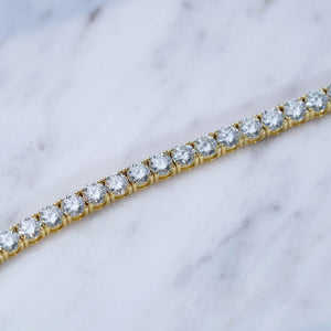 Horizontal close-up detail shot of 4mm round brilliant D color VVS1 clarity moissanite tennis bracelet, solid S925 sterling silver core with thick 18K yellow gold plating, each clear moissanite gem hand prong set with polished gold prongs, placed on clean white grey marble texture surface, bright natural lighting highlighting outstanding sparkle, fire and clarity of flawless moissanite stones, unisex hip hop iced out stackable wrist jewelry for men and women, tarnish resistant daily wear luxury tennis chain