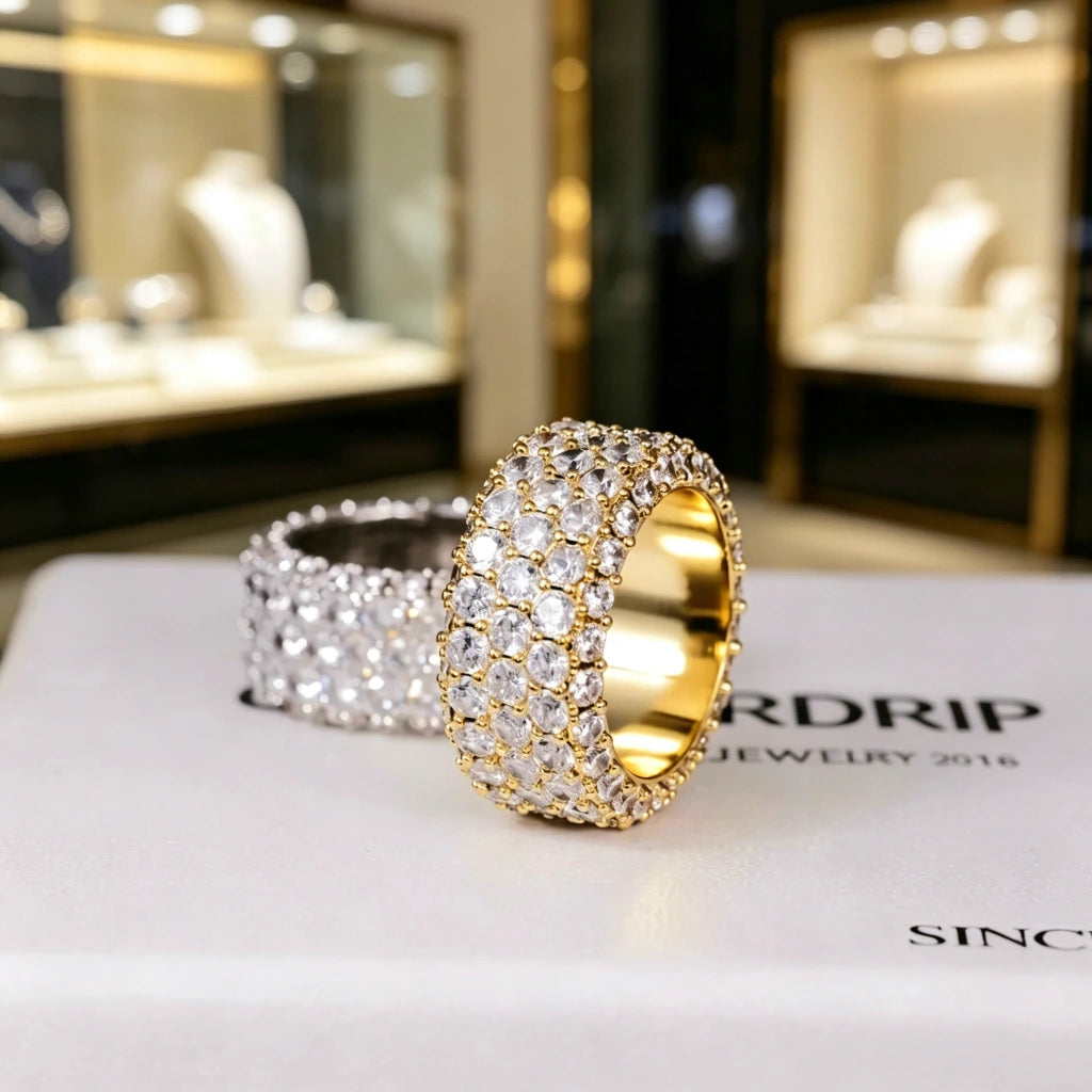 RDRIP JEWELRY 5‑Row Fully Paved Iced Out Eternity Ring displayed on branded RDRIP JEWELRY SINCE 2016 box, gold‑toned prong‑set ring in foreground with silver‑toned ring blurred in background, heavy 14mm wide hip hop eternity band ring, full‑around hand‑set flawless VVS simulated diamonds, premium 18K & white gold plated icy bling hip‑hop jewelry for men and women, Prong Cuban family statement ring against soft bokeh jewelry store showcase window background

