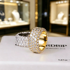 RDRIP JEWELRY 5‑Row Fully Paved Iced Out Eternity Ring displayed on branded RDRIP JEWELRY SINCE 2016 box, gold‑toned prong‑set ring in foreground with silver‑toned ring blurred in background, heavy 14mm wide hip hop eternity band ring, full‑around hand‑set flawless VVS simulated diamonds, premium 18K & white gold plated icy bling hip‑hop jewelry for men and women, Prong Cuban family statement ring against soft bokeh jewelry store showcase window background
