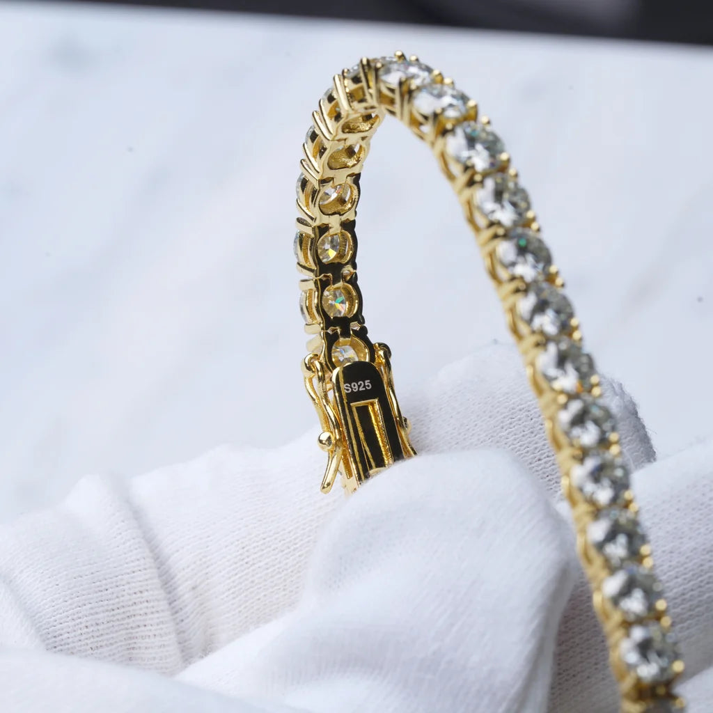 Detailed curved close-up shot of 5mm Hearts & Arrows cut D Color VVS1+ moissanite tennis chain with thick 18K yellow gold plating over solid S925 sterling silver, dual safety lock clasp clearly stamped S925 on inner metal surface, each colorless flawless moissanite stone hand prong-set by professional artisans showing vivid rainbow fire and bright crystal shine, bent golden wrist chain gently held by soft clean white cotton jewelry gloves, faint light marble texture as blurred background, reinforced double 