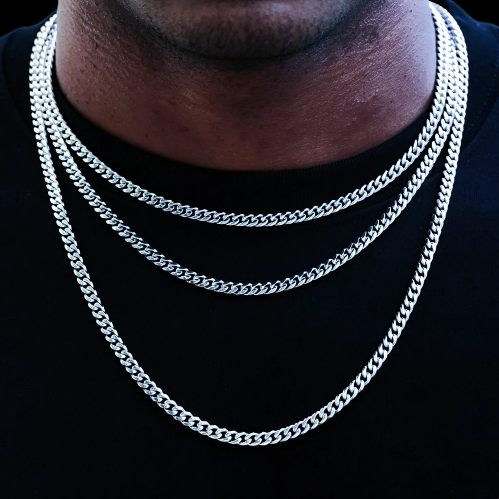 Studio shot with solid pure black background, black male model wearing layered stackable 5mm 18k white gold Miami Cuban link chain set, three multi-length cuban necklaces layered for hip hop streetwear styling, smooth fully polished tangle-free flexible uniform links, premium white gold plated anti-tarnish long-lasting shiny men’s urban statement jewelry, versatile design to wear alone or pair with custom hip hop pendants, expert hand-finished handcrafted artisan cuban chain, suitable for daily casual
