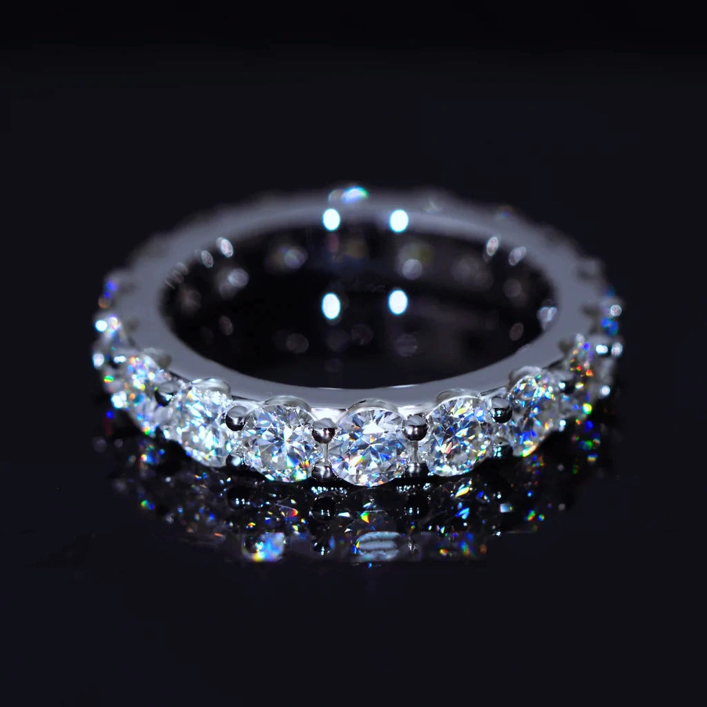 6.00 CARAT S925 VVS1+ moissanite single‑row full eternity ring, 4mm round ideal cut hearts and arrows moissanite stones, white gold plated genuine 925 sterling silver band, brilliant rainbow dispersion light reflection, full band ring resting on dark glossy black reflective surface, low‑light studio close‑up shot, icy sparkling unisex hip‑hop wedding stackable fine bling jewelry RDRIP JEWELRY