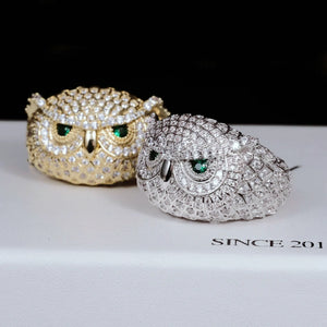 Depth of field product photograph of Iced Owl King Ring, foreground white gold plated owl ring and background 18K gold plated owl ring, realistic feather scale detailing, glowing emerald green eyes, fully encrusted flawless VVS simulated diamonds, heavy hip hop men bling jewelry laid on branded display box RDRIP JEWELRY
