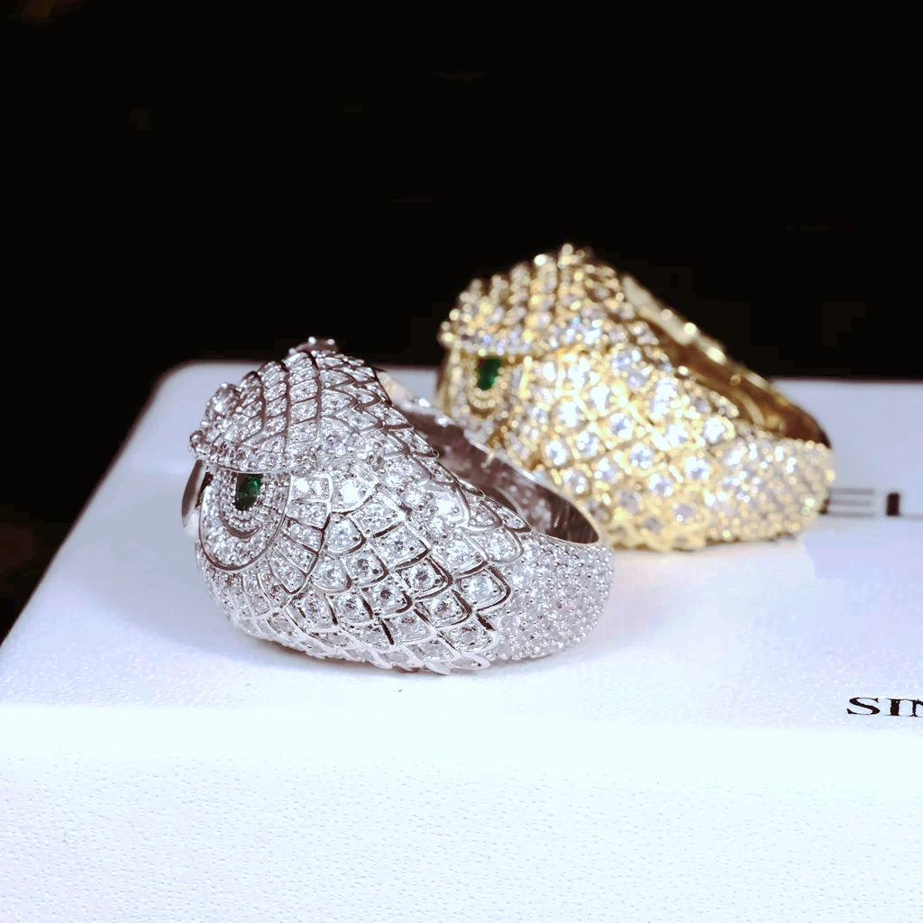 Side profile angle shot of Iced Owl King Ring, white gold plated owl ring in foreground showing band inner detail, 18K gold plated owl ring softly blurred in background, intricate owl feather scale carvings, vivid emerald green eyes, fully encrusted flawless VVS simulated diamonds, premium heavy hip hop mens bling ring on white display surface RDRIP JEWELRY
