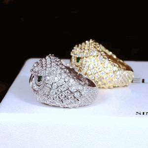 Side profile angle shot of Iced Owl King Ring, white gold plated owl ring in foreground showing band inner detail, 18K gold plated owl ring softly blurred in background, intricate owl feather scale carvings, vivid emerald green eyes, fully encrusted flawless VVS simulated diamonds, premium heavy hip hop mens bling ring on white display surface RDRIP JEWELRY
