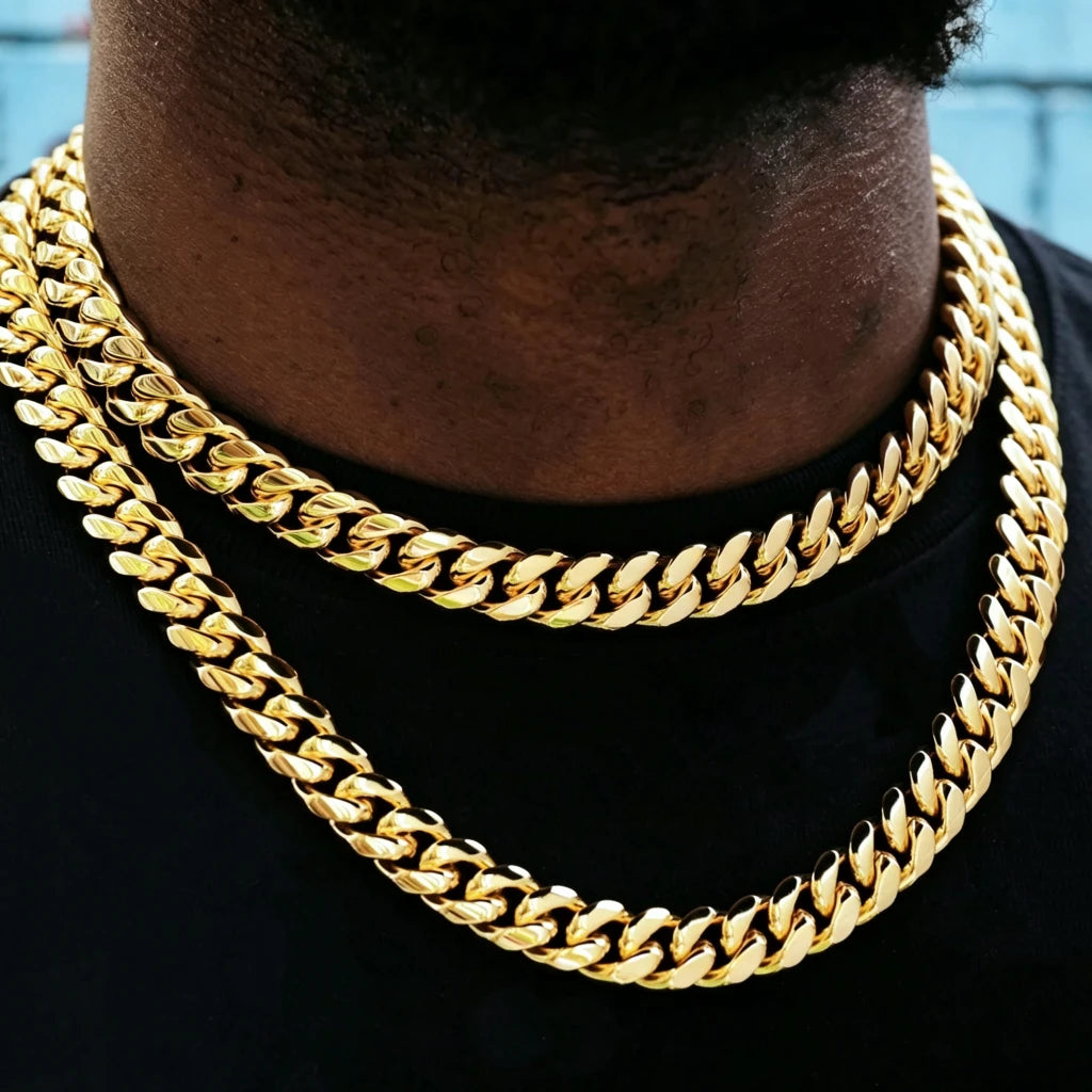 Extreme close-up shot of 12.2mm wide 18k gold plated Miami Cuban link choker with sturdy box clasp, two layered stacked heavy curb gold chains worn on Black male neck, soft minimal light blue wall background with subtle faint street graffiti details, tight polished interlocking cuban links, bright reflective anti-tarnish gold finish, thick hip hop layering necklace for men streetwear, bold icy luxury street jewelry closeup product photo, stackable gold curb chain everyday fashion accessory RDRIP JEWELRY