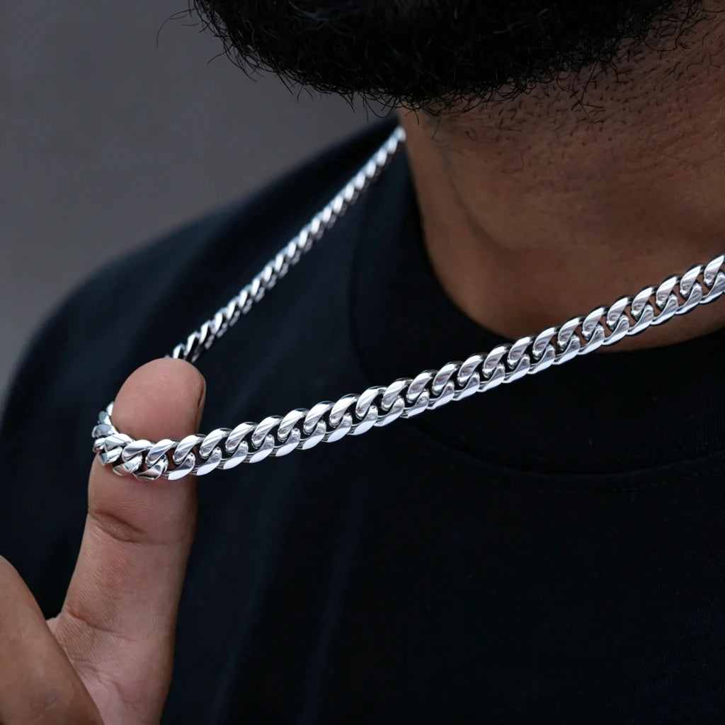 Extreme closeup shot of a black man's finger holding polished 8mm Flagship Miami Cuban Link Chain in White Gold Tone, tight interlock stainless steel curb necklace with bright icy silver shine, solid dark blurred background, smooth scratch-resistant anti-tarnish metal links, logo-free blank clasp design, versatile men hip hop streetwear jewelry for single wear or layered stacking, available in 16 18 20 22 24 inch lengths, heavy durable everyday fashion accessory, complete gift set with storage pouch and lif