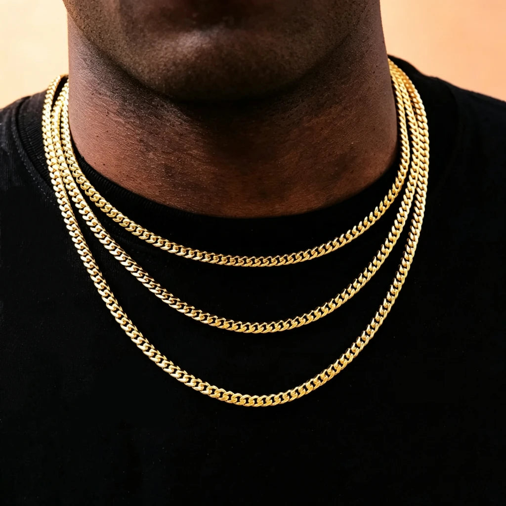 Front close-up portrait of black male model with trimmed short stubble beard wearing three layered stackable 5mm Miami Cuban Link Chain in 18k gold plated, polished glossy solid cuban link necklace for men hip hop streetwear, shot on soft light peach background, flexible tangle-free construction, available lengths 18 20 24 30 inch with weight tolerance ±5 grams, anti-tarnish durable 18k gold plating, can be worn alone or matched with custom hip hop pendants, classic urban layering jewelry gift for men