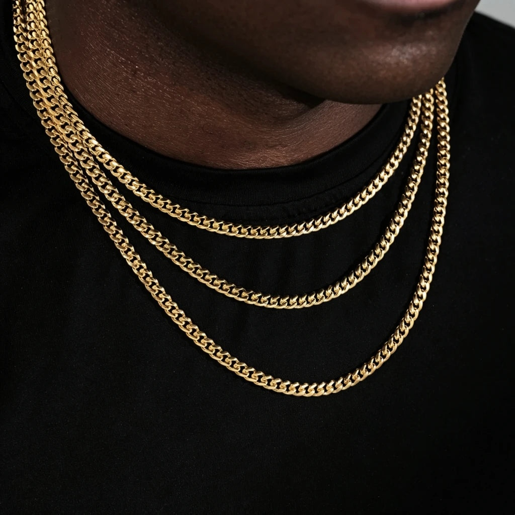 Close cropped jawline shot of Black male model with trimmed short stubble, showcasing three layered stackable 5mm Miami Cuban Link Chain in 18k gold plated, polished glossy gold cuban necklace for men hip hop streetwear, solid flexible tangle-free link construction, available sizes 18, 20, 24, 30 inch with weight range 24g-38g and ±5g tolerance, long-lasting anti-tarnish 18k gold plating, versatile layering chain that can be worn alone or matched with custom hip hop pendants, minimalist black tee backdrop