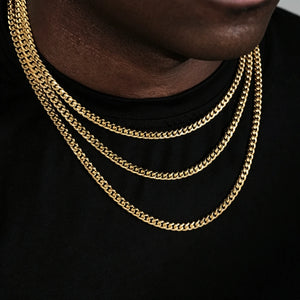 Close cropped jawline shot of Black male model with trimmed short stubble, showcasing three layered stackable 5mm Miami Cuban Link Chain in 18k gold plated, polished glossy gold cuban necklace for men hip hop streetwear, solid flexible tangle-free link construction, available sizes 18, 20, 24, 30 inch with weight range 24g-38g and ±5g tolerance, long-lasting anti-tarnish 18k gold plating, versatile layering chain that can be worn alone or matched with custom hip hop pendants, minimalist black tee backdrop