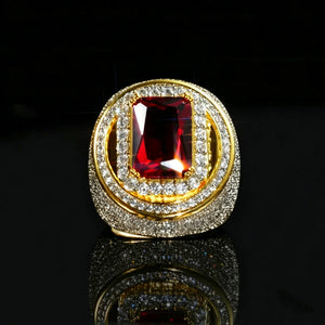 Front halo close‑up studio shot of two‑tone 18k gold plated men hip‑hop champion ring, eye‑catching large radiant‑cut red ruby center gemstone, double layered halo setting with hand‑set micro pave cubic zirconia stones, heavy thick wide band, brilliant icy bling sparkle, resting on glossy black reflective mirror surface, deep dark background, premium bold rapper statement fashion jewelry RDRIP JEWELRY
