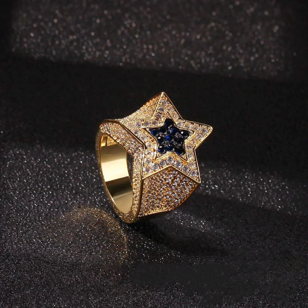 Tilted angle shot of men’s gold plated iced out double pentagram star hip hop ring, visible polished inner band, full micro pave AAA white cubic zirconia across wide heavy band, center inlaid deep royal blue CZ star, hand‑set bling statement rap jewelry for street fashion, night party and holiday gift for him from RDRIP JEWELRY

