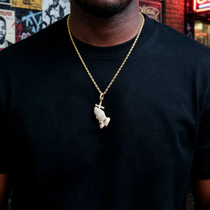 Black male model wearing gold iced out praying hands cross pendant necklace with gold twisted rope chain, 18k gold plated vvs simulated diamonds hip hop faith jewelry, urban american graffiti street wall background men streetwear bling shot RDRIP JEWELRY
