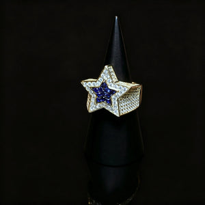 Men’s gold plated iced out double pentagram star hip‑hop ring displayed on black cone ring stand, hand‑set AAA white cubic zirconia surrounding vivid deep‑blue CZ star center, heavy wide micro pave band bling statement ring, premium rap streetwear jewelry piece for men party and gift giving from RDRIP JEWELRY
