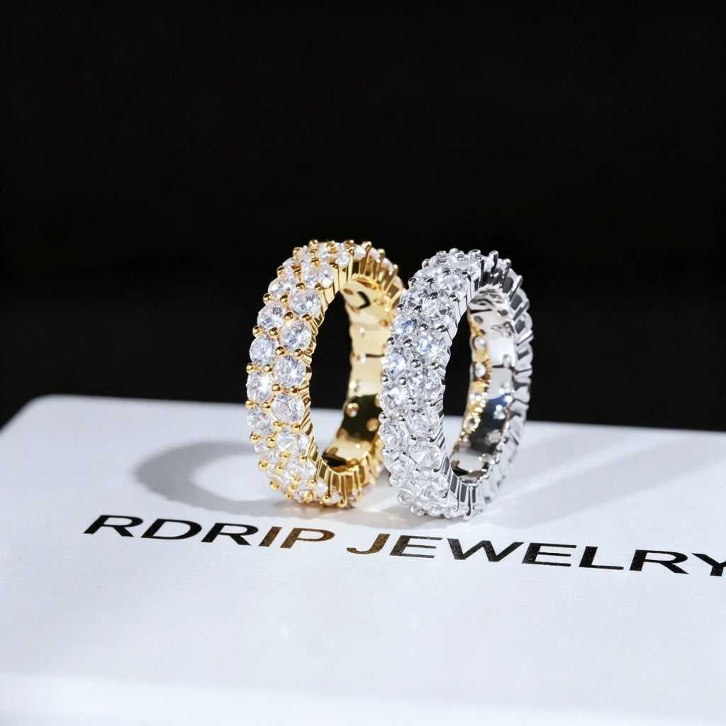 ```
RDRIP JEWELRY iced out double row eternity band gold silver pair resting on branded white gift box prong‑set 3mm round VVS simulated diamonds full iced hip hop mens stackable ring 6mm width copper base bling jewelry dark background studio product close‑up shot