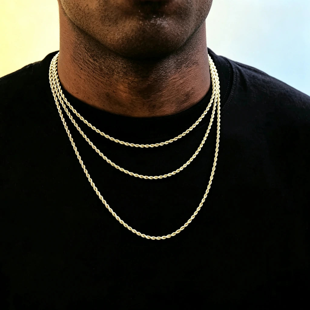 Medium full studio shot of African American male model with ultra short clean skin stubble beard, wearing three different length stackable 2mm 18k gold plated twisted rope chain layered necklaces, slim dainty twisted link staple jewelry for hip hop streetwear men, minimalist unisex gold chain that can be worn alone or matched with custom brand pendants, hand-finished anti-tarnish long-lasting shiny rope chain, smooth soft pale yellow and light blue gradient solid background, clean commercial product 