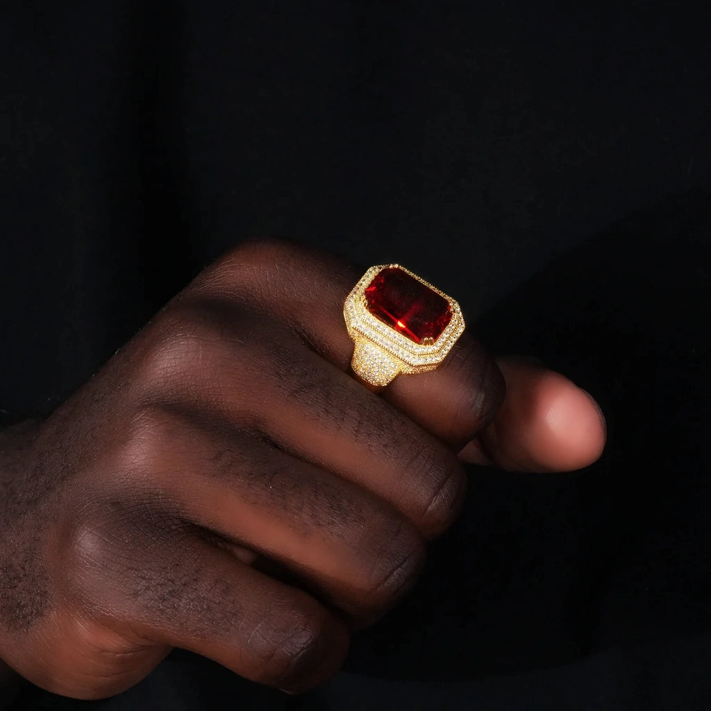 Man wearing heavy iced‑out men’s hip‑hop ring on hand, large emerald‑cut deep red gemstone center, double halo hand‑set micro‑pave sparkling stones, chunky wide 18K gold plated band, bold urban hip hop fashion jewelry, real hand model wearing shot against dark background RDRIP JEWELRY

