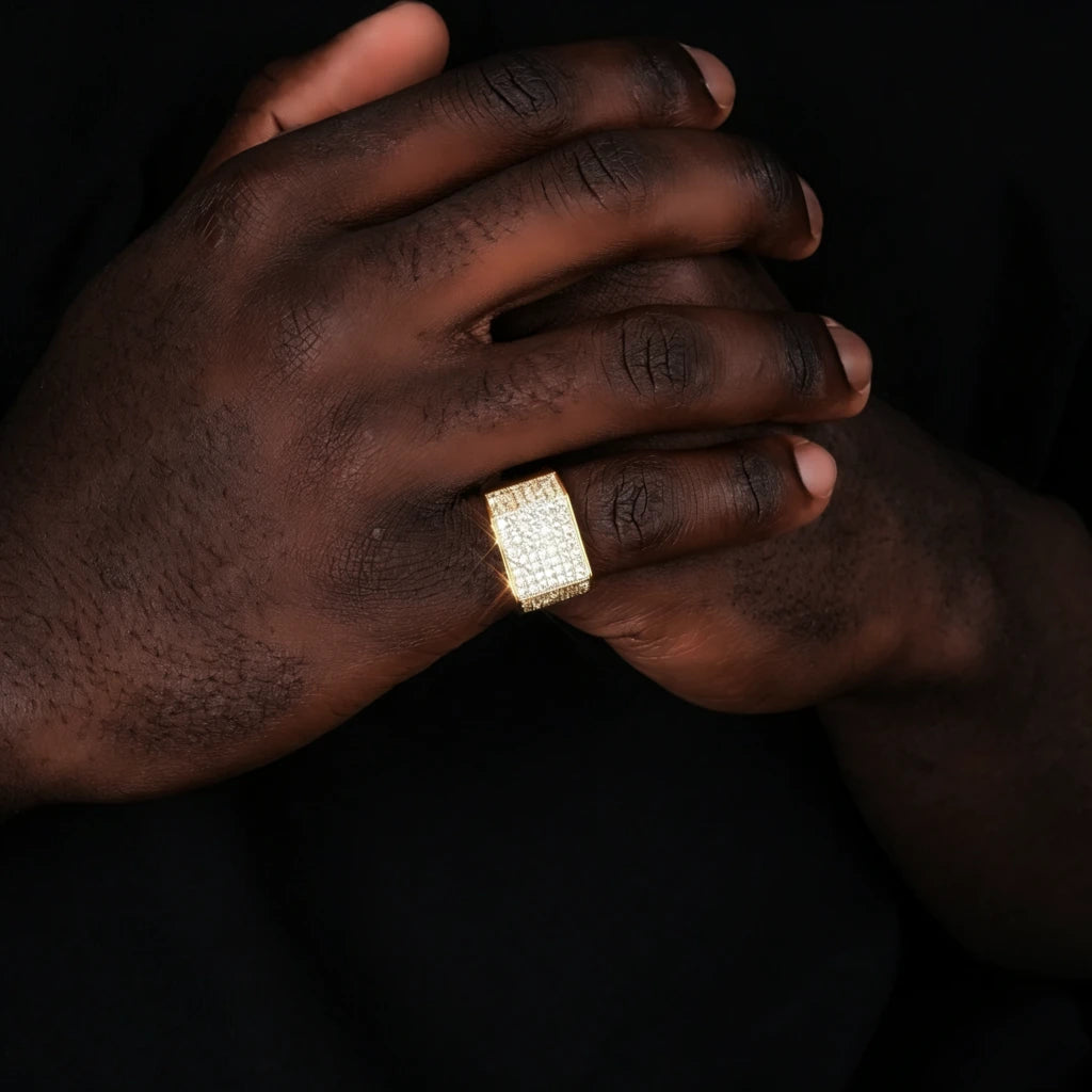 Black male model wearing two‑tone gold silver full pave square statement band ring from RDRIP JEWELRY, men iced‑out bling hip hop ring on dark skin hand, wide square face densely hand‑set cubic zirconia and rhinestone stones, 18k gold plated copper heavy statement jewelry, real‑life hand wearing shot against solid black background
