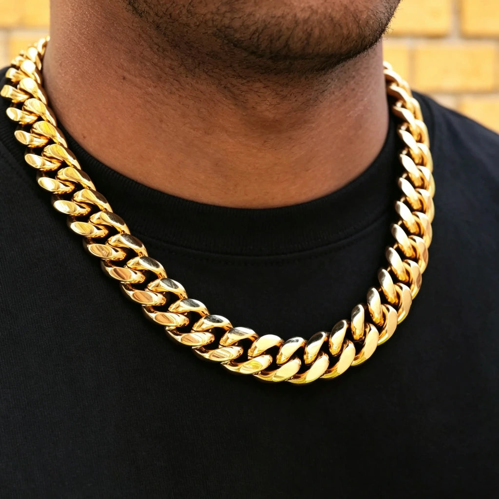 Upper body shot of male model wearing oversized 18mm chunky yellow gold plated Miami Cuban link choker necklace, heavy thick polished curb hip hop chain, neatly trimmed short beard, spotless plain solid black crew neck tee with zero lint or dust, warm textured yellow brick wall background, mirror shiny reflective gold finish, tight interlocking solid links heavyweight men streetwear statement jewelry, stackable hip-hop layering accessory with secure box clasp, anti-tarnish gold plated everyday wear luxury s