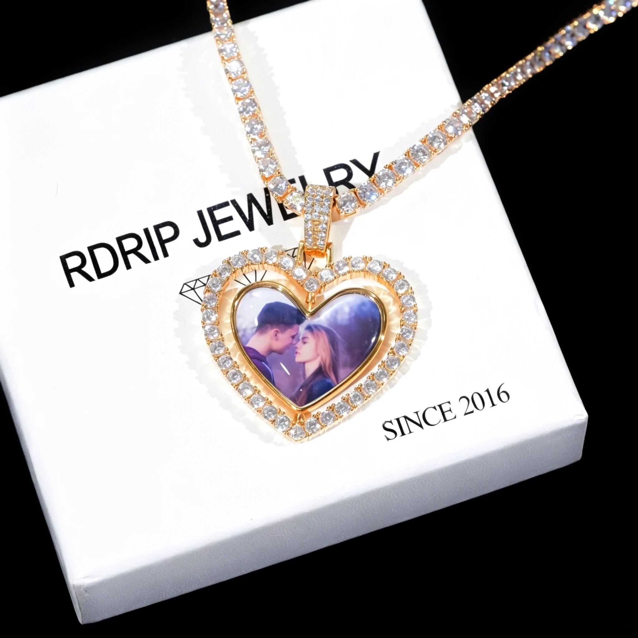 Iced Out Double Sided Rotatable Heart Custom Photo Pendant, gold tone with tennis chain, couple portrait inside heart frame, placed on white RDRIP JEWELRY branded box printed SINCE 2016, dark textured background personalized photo locket necklace, RDRIP JEWELRY
