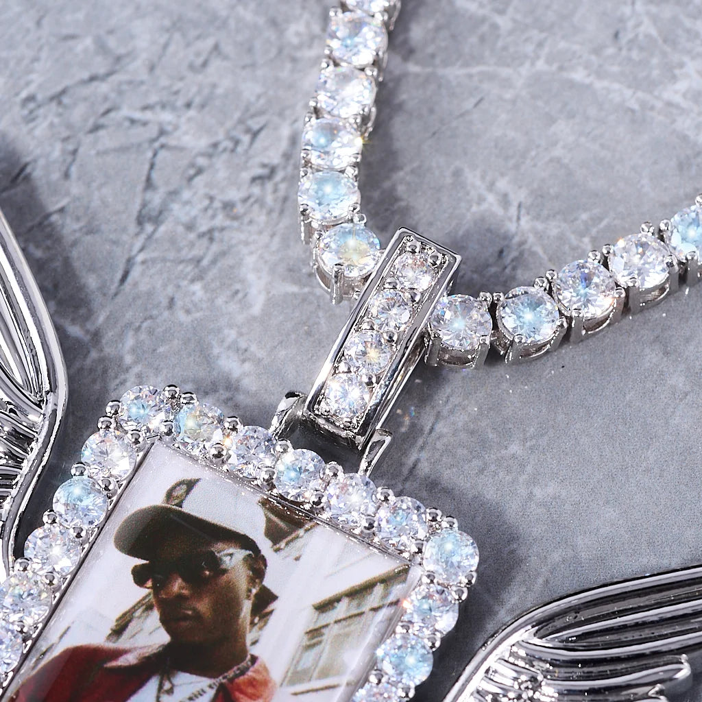 Iced Out Wings RIP Custom Photo Pendant, white gold tone close-up detail shot, focus on encrusted stone frame, custom photo insert and fully iced bail, partial angel wings & matching tennis chain, grey marble texture background, memorial personalized hip hop photo jewelry detail view, RDRIP JEWELRY
