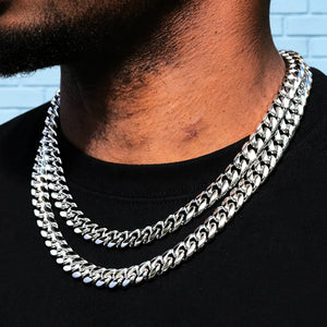 Outdoor side profile close-up shot of Black male wearing double layered 12.2mm non-stone 14k white gold plated Miami Cuban link chain with sturdy box lock clasp, neatly trimmed short beard, simple black crew neck t-shirt, soft pale light blue textured brick wall background, thick tightly interlocking polished curb links, bright cool silver-white anti-tarnish plating, heavy weight plain uniced hip hop stackable necklace for men streetwear layering, premium minimalist men bling jewelry lifestyle photo RDRIP 