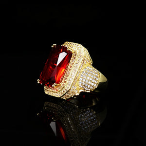 Side angle product shot of men’s heavy iced out hip‑hop statement ring, large emerald‑cut deep red gemstone secured by gold prongs, full hand‑set micro pave double halo setting, densely encrusted wide ring shank, premium 18K gold plated finish, ornate chunky hip hop men jewelry, displayed on glossy black reflective surface with mirror reflection RDRIP JEWELRY

