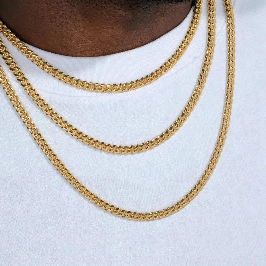 Product display close-up shot of three layered stackable 5mm Miami Cuban Link Chain with polished 18k gold plated finish, fitted on dark brown Black male neck mannequin wearing plain clean white crew neck t-shirt, shiny reflective gold cuban links with smooth flexible tangle-free construction, available necklace lengths 18 inch, 20 inch, 24 inch, 30 inch, long-lasting anti-tarnish gold plating resistant to fading and oxidation, versatile stackable layering chain that can be worn alone or matched with custom